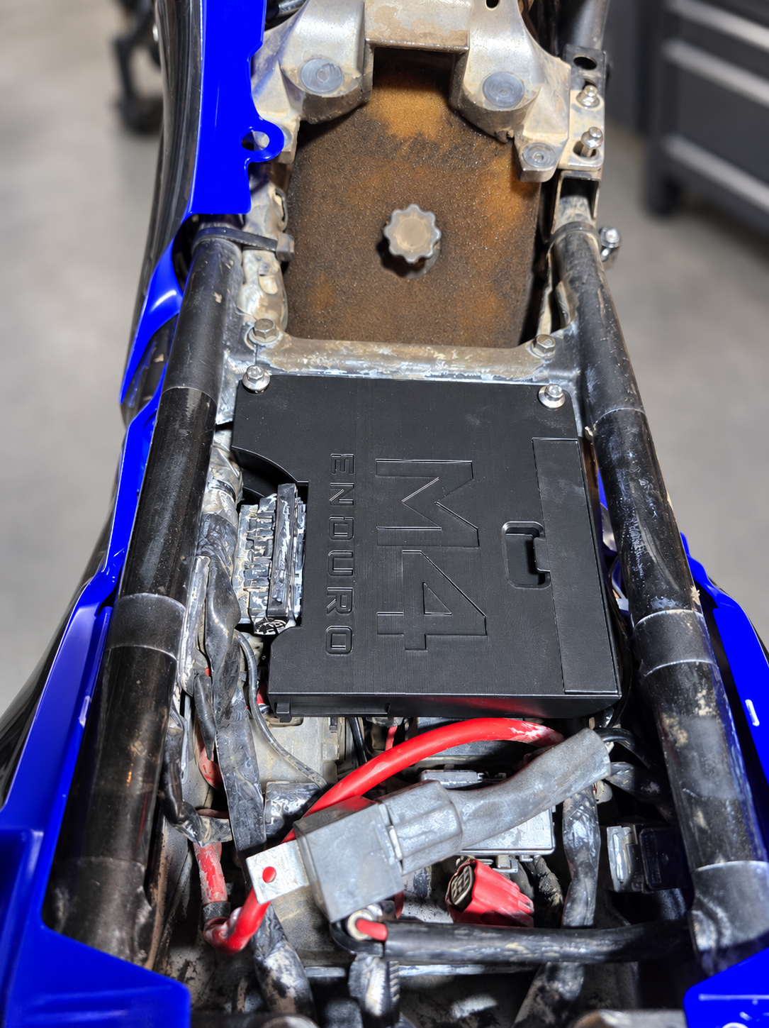 Top view of the M4 Enduro ECU relocate kit fitted between the Yamaha Ténéré 700 frame rails beneath the seat, with the garage background cleaned and softly blurred.